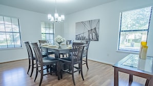 A dining area with a round table, six chairs, a chandelier, and a cityscape painting on the wall.