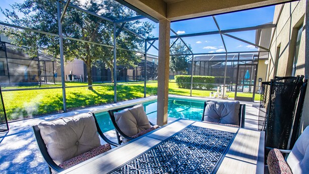A covered outdoor seating area with a pool view, featuring a table and chairs with patterned cushions.