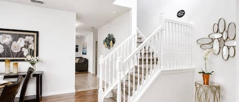 Una escalera con barandillas blancas que lleva a un piso superior, un comedor con mesa y sillas, y un espejo adosado a la pared con detalles decorativos.