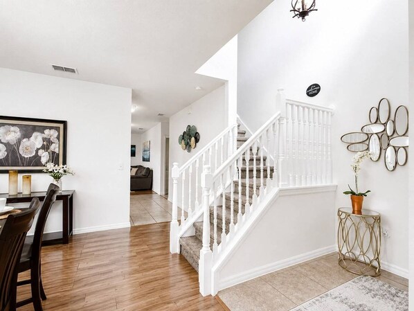 Una escalera con barandillas blancas que lleva a un piso superior, un comedor con mesa y sillas, y un espejo adosado a la pared con detalles decorativos.