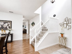 A staircase with white railings leading to an upper floor, a dining area with a table and chairs, and a wall-mounted mirror with decorative elements.