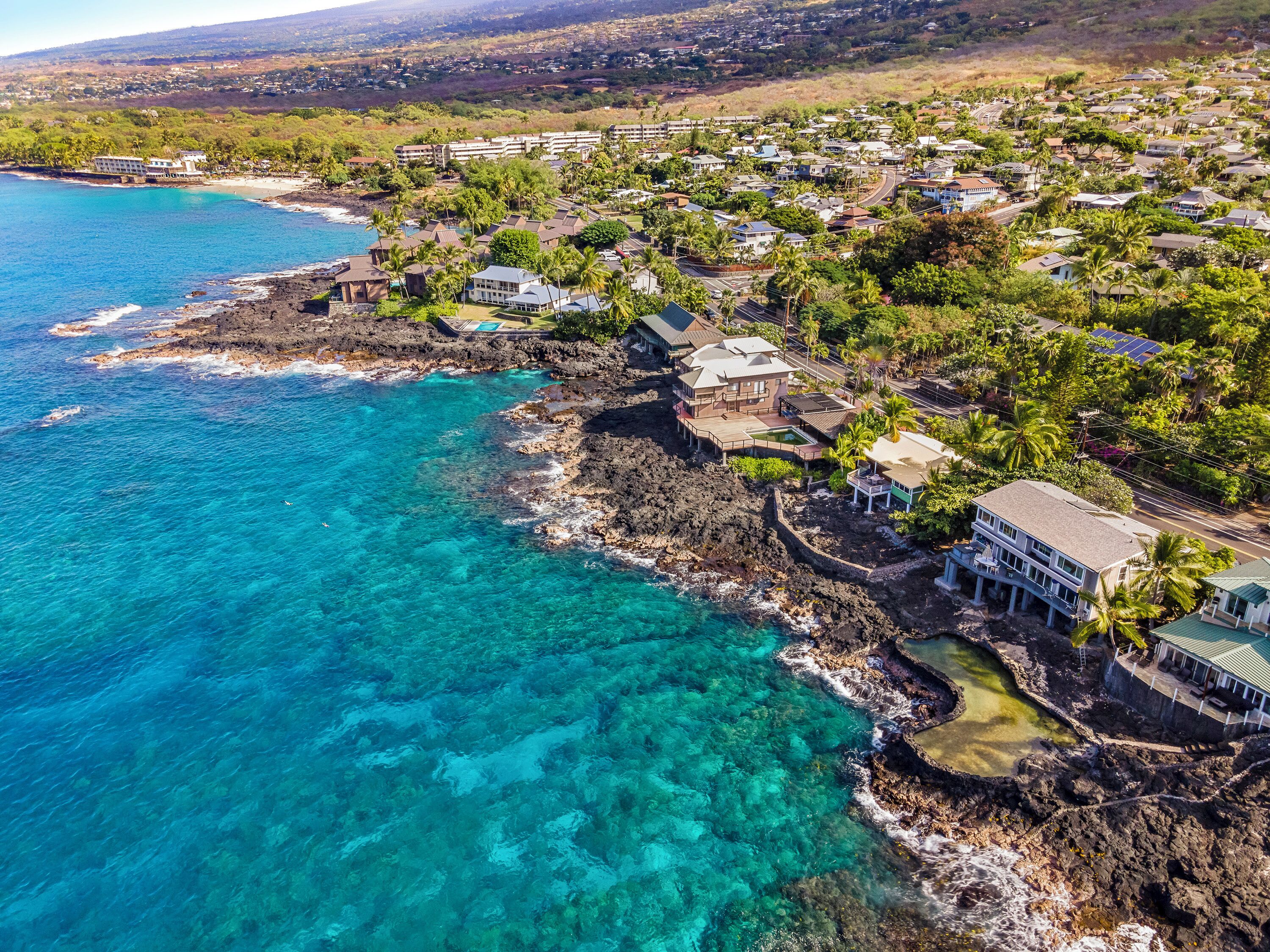 An aerial view of a coastal town with houses, clear blue waters, and a rocky shoreline.