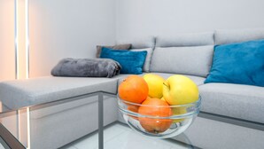 A modern living room with a glass coffee table holding fruit, a grey sofa with blue cushions, and a grey throw blanket.