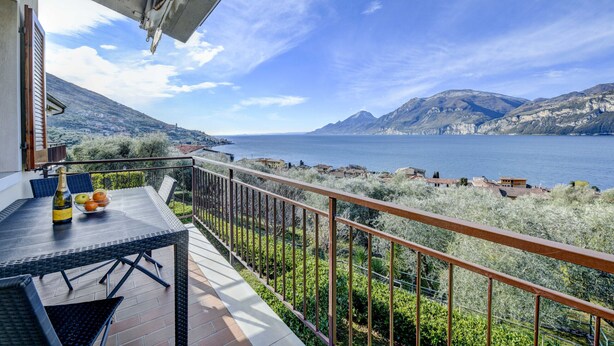 A balcony with a table, chairs, and a bottle of champagne. The balcony overlooks a scenic view of a lake and mountains.