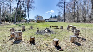Un campamento con un hoyo para fuego rodeado de troncos de madera, un campo de césped y casas al fondo.