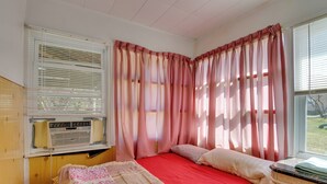 Un dormitorio con una cama roja, una ventana con persianas, un calentador y una mesita de noche con una lámpara.