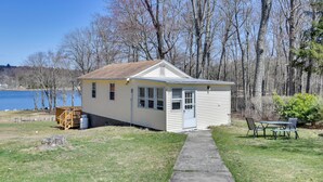 A small, single-story house with a front porch and a visible garage door, situated next to a body of water and surrounded by trees. - Shared Fire Pit & Kayaks: Catskills Lake Retreat! (Ellenville)
