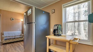 A compact kitchen with a refrigerator, a coffee maker, a wooden table, a sofa, and a window with a view of trees. - Shared Fire Pit & Kayaks: Catskills Lake Retreat! (Ellenville)