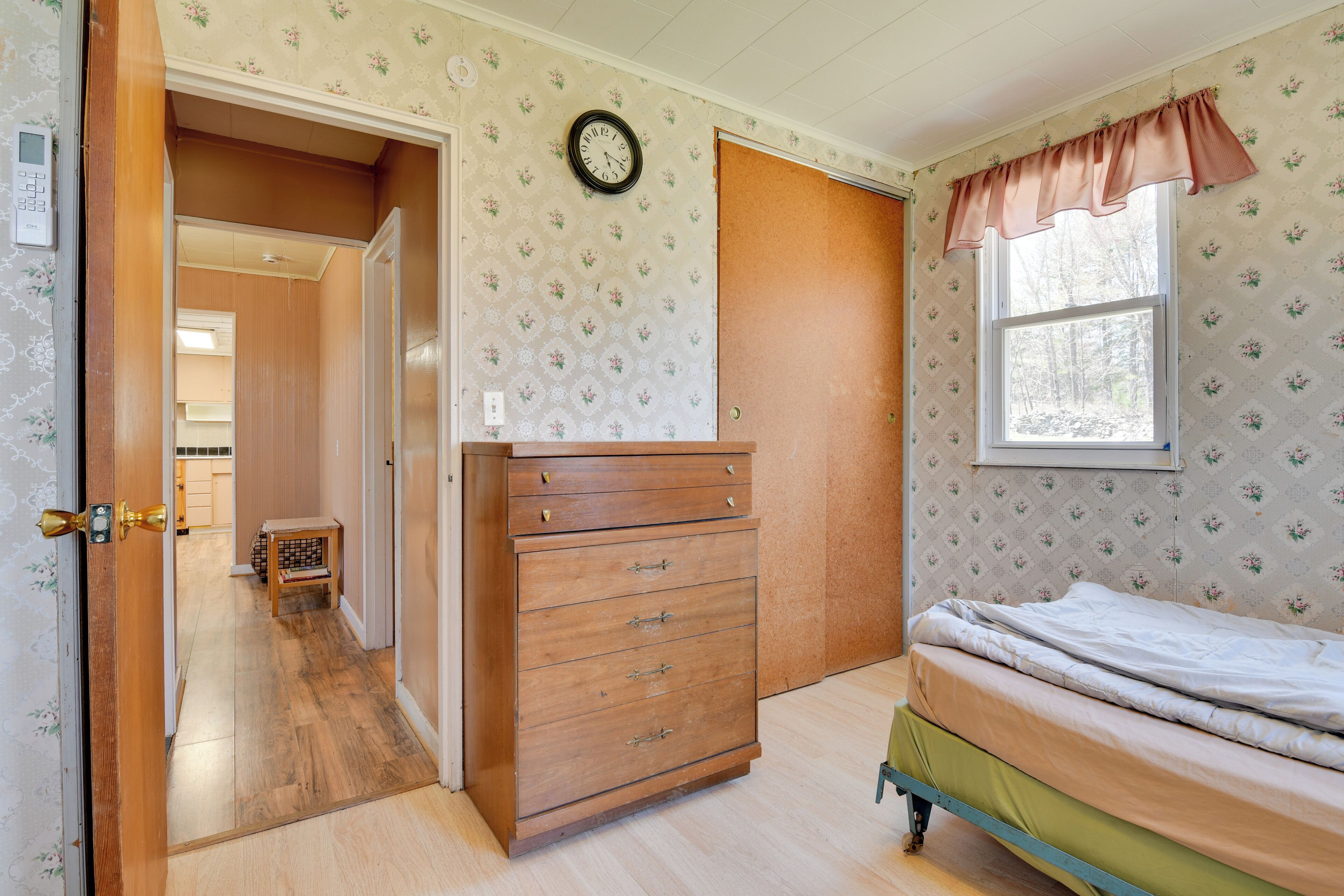 Ein Schlafzimmer mit einem hölzernen Schminktisch, einem Bett mit grüner Bettdecke, einem Fenster mit floralem Vorhang und einer Uhr an der Wand.