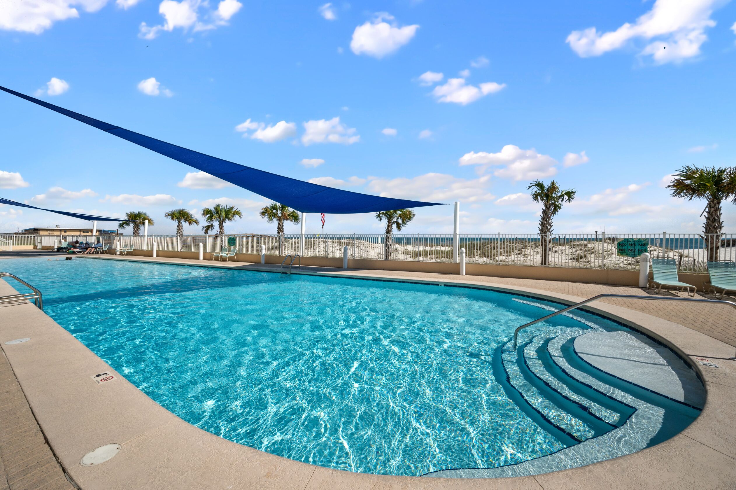 A clear blue pool with a shaded area, palm trees, and a fence.