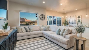 A modern living room with a large sectional sofa, a large window with a desert view, a clock on the wall, and a chandelier. - O-29&30| 2 Homes! Private Pool & Hot Tubs * Views (Santa Clara)