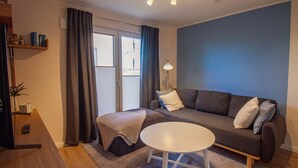 Una sala de estar moderna con un sofá gris, una mesa de centro blanca y una lámpara. Hay una ventana con cortinas, un estante con adornos y una mesita de noche de madera.