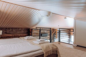 Un dormitorio con techo de madera, una cama con sábanas blancas, una manta y vista a una cabaña de troncos a través de una ventana.