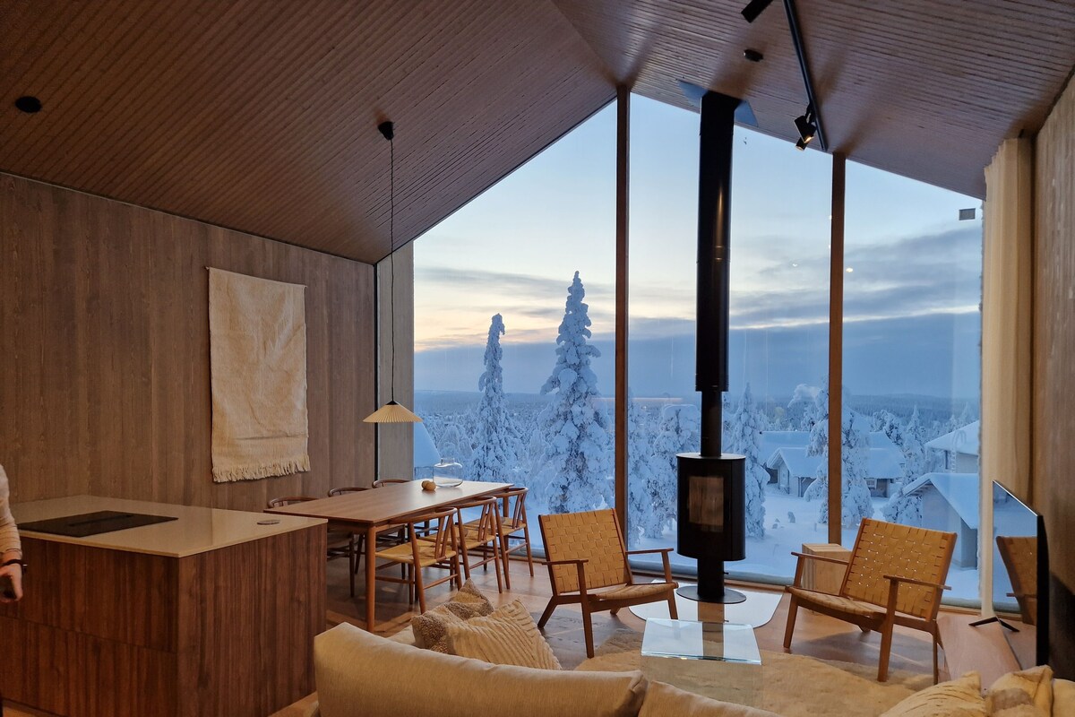 Un intérieur moderne avec un plafond en bois, une grande fenêtre donnant sur un paysage enneigé, une cheminée et un coin salon confortable.