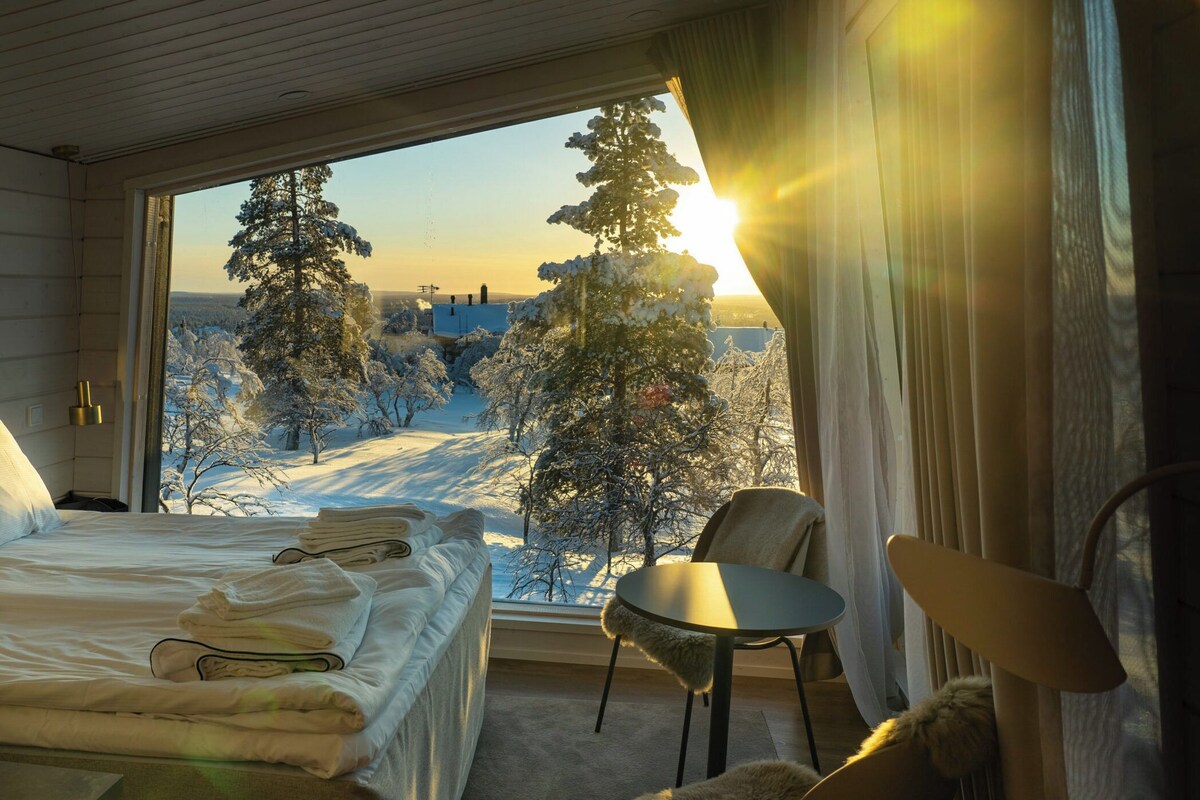 Une chambre d’hôtel avec un lit, une chaise, une table et une vue sur un paysage enneigé bordé d’arbres.