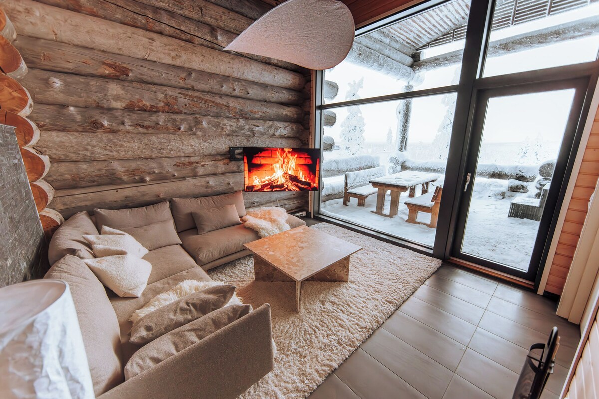 L’intérieur d’un chalet confortable, avec une cheminée, un canapé, une table basse et une grande fenêtre donnant sur un paysage enneigé.