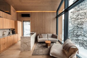 Un interior moderno con paneles de madera, una zona de cocina, un sofá y un ventanal con vistas nevadas.