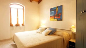 A bedroom with a bed, a window, a painting, and a nightstand with a lamp.