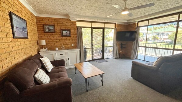 A living room with a brick wall, a sofa, a coffee table, a TV, and a sliding glass door leading to an outdoor area. - Seacrest 6 - walk to the beach - Bring your own linen (SOUTH WEST ROCKS)