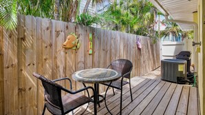 A wooden deck with a round table and two chairs, a wooden fence with a decorative fish head, and a white building in the background. - Sand and Sea 109 A! Pool, Patio and Coastal Charm! (Anna Maria)