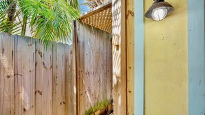 A wooden fence with a small opening, a wooden deck, and a wall with a light fixture. - Quaint Condo with Shared Pool, Only 2 Houses to the Beach - Sand And Sea 109 B (Anna Maria)