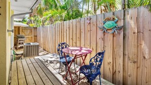 A wooden deck with a table and chairs, a wooden fence, and a decorative crab. - Quaint Condo with Shared Pool, Only 2 Houses to the Beach - Sand And Sea 109 B (Anna Maria)