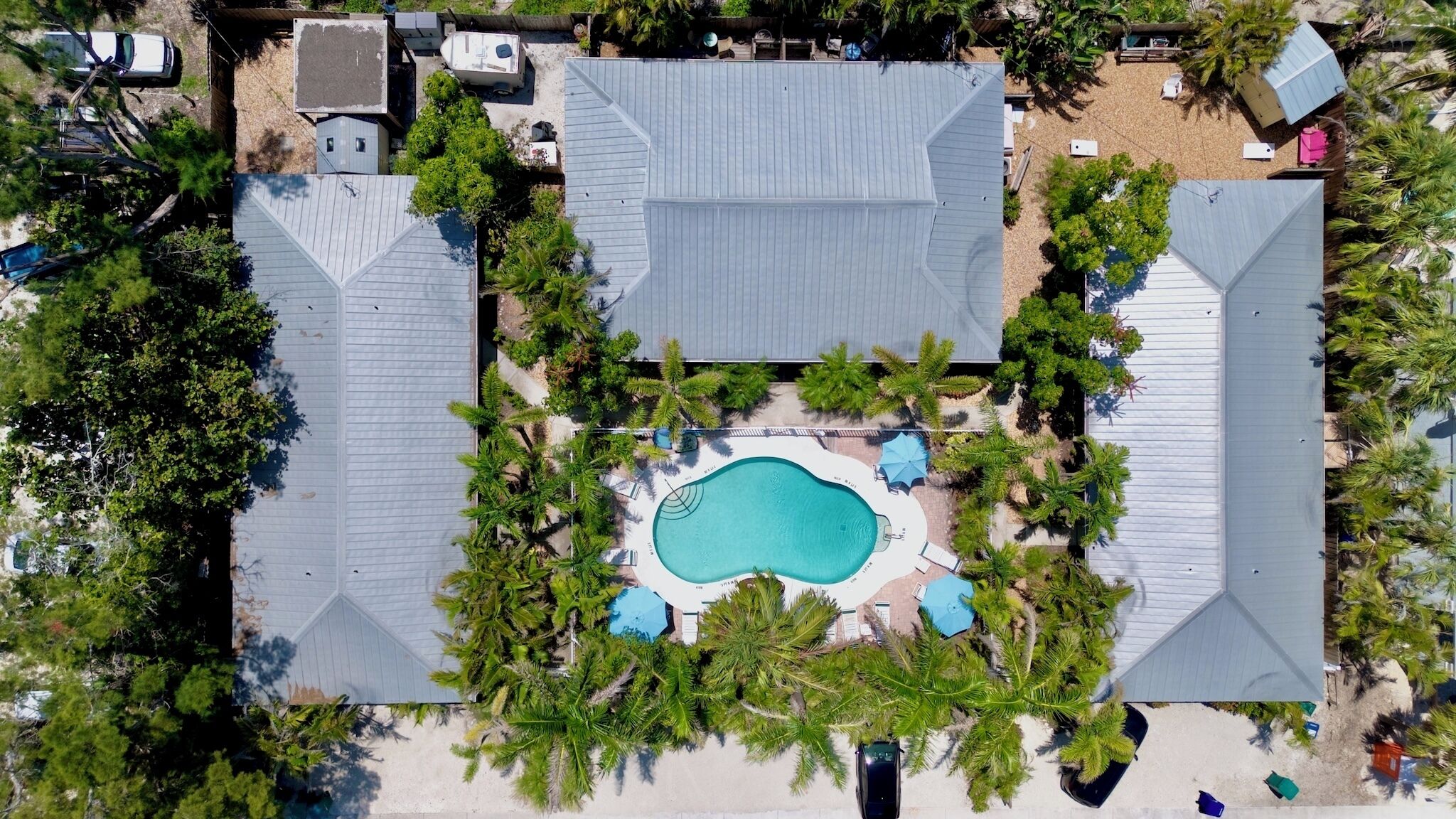 Vue aérienne d’un complexe hôtelier doté d’une piscine centrale, entourée de palmiers et de structures abritées.