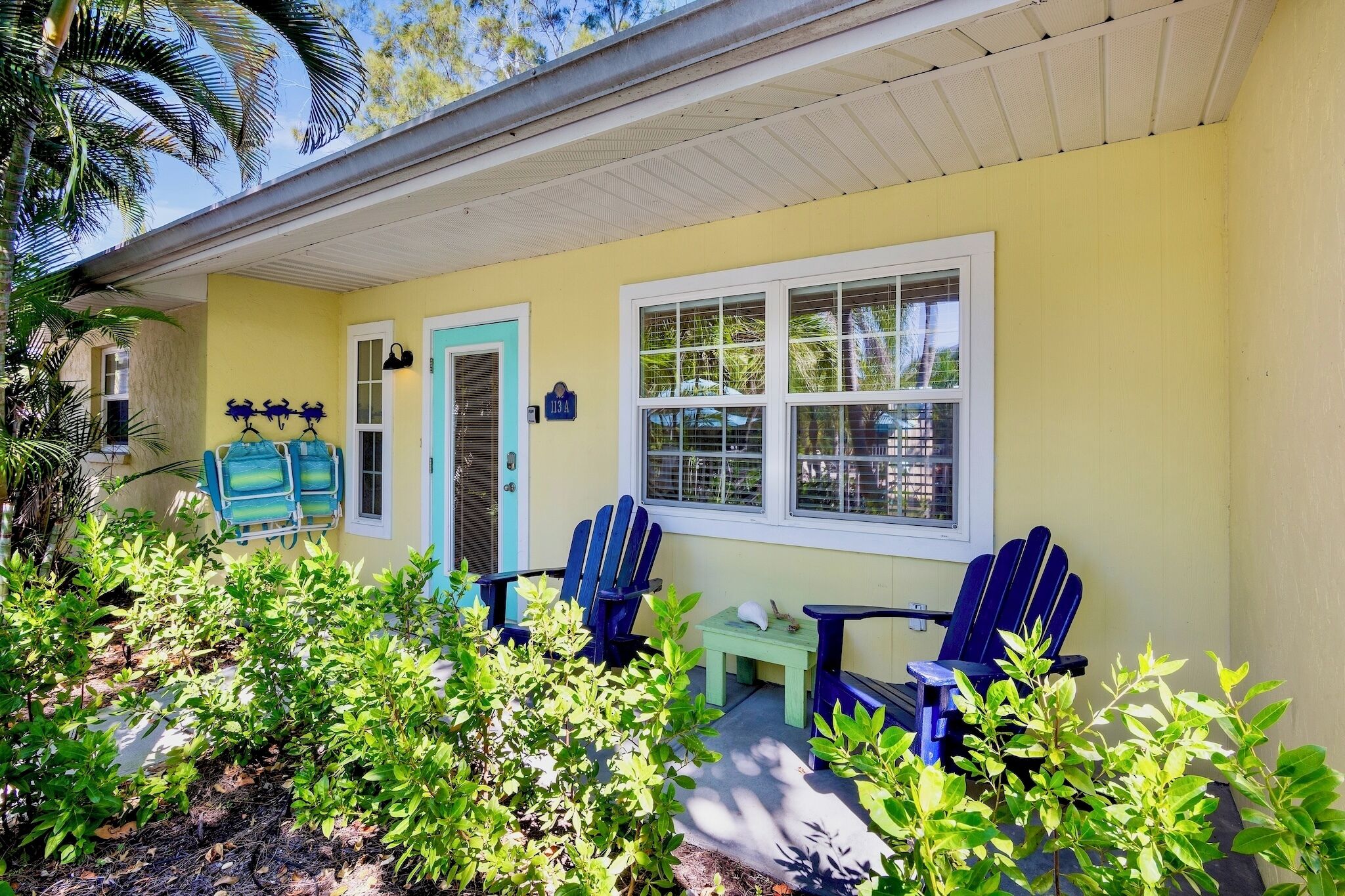 Una casa amarilla con porche, sillas Adirondack azules y una pequeña mesa.
