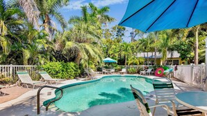 A pool area with lounge chairs, a table, and a blue umbrella. - Steps to Beach and Pool! Cozy Island Flat Near Pine Ave! (Anna Maria)