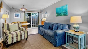 A living room with a striped armchair, a blue sofa, and a small table with a lamp. There is a painting on the wall and a window with curtains. - Steps to Beach and Pool! Cozy Island Flat Near Pine Ave! (Anna Maria)