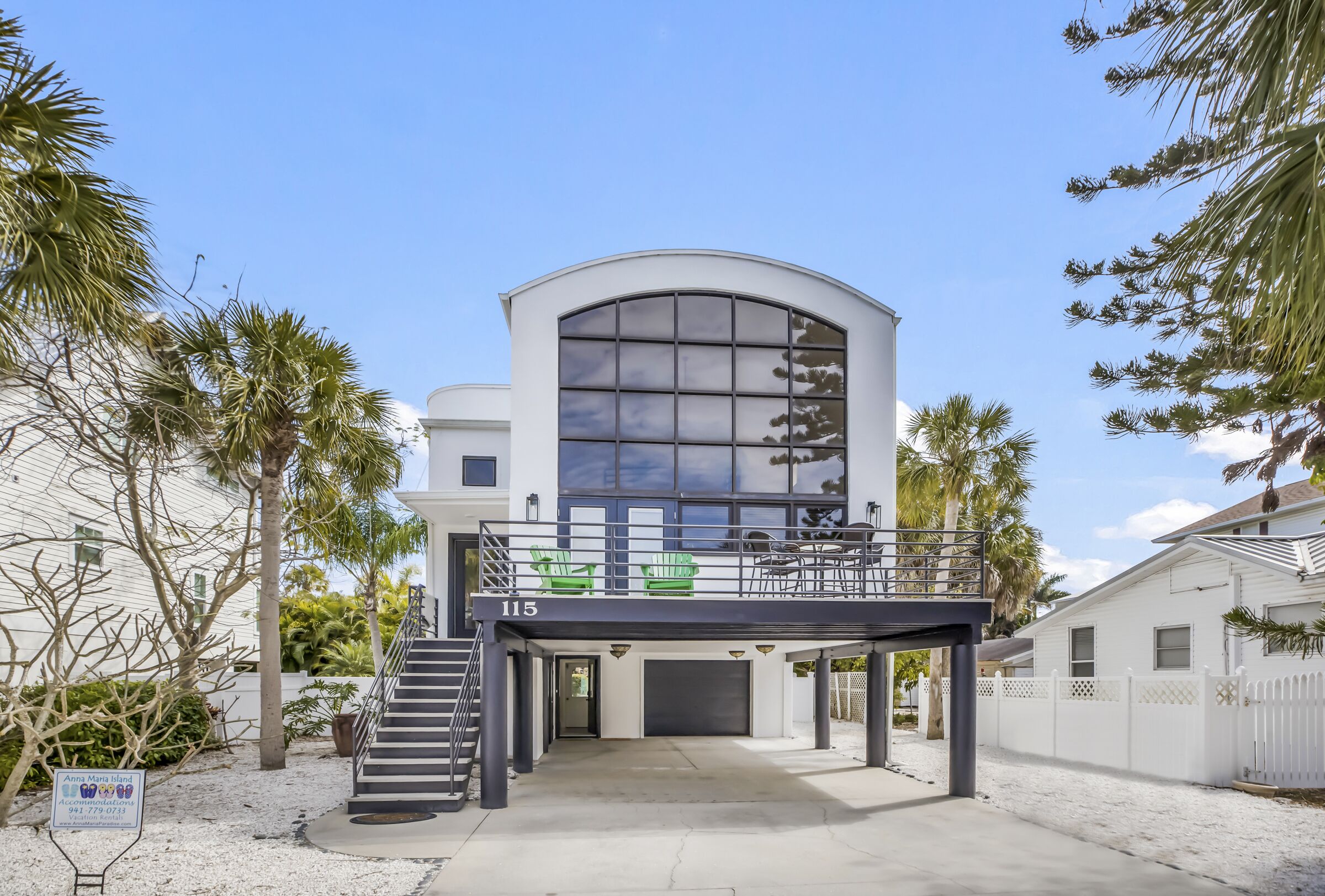 A modern two-story building with a curved glass facade, a covered outdoor seating area with green chairs, and a sign indicating it is Anna Maria Island.