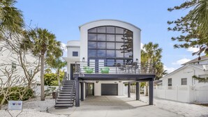 A modern two-story building with a curved glass facade, a covered outdoor seating area with green chairs, and a sign indicating it is Anna Maria Island. - Luxury home near the beach and shopping! Private backyard and pool! (Anna Maria)