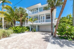 A two-story beachfront property with a covered driveway, palm trees, and a clear blue sky.