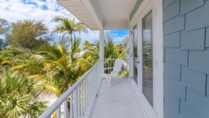 A covered porch with a white railing, a single white chair, and a view of palm trees and a house in the background.