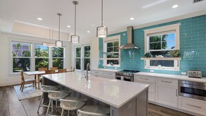 A modern kitchen with a large island, bar stools, and a view of the outdoors through large windows.