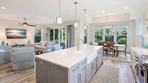 A modern kitchen with a central island, a dining area with wooden chairs, and a living room with a sofa and a TV.