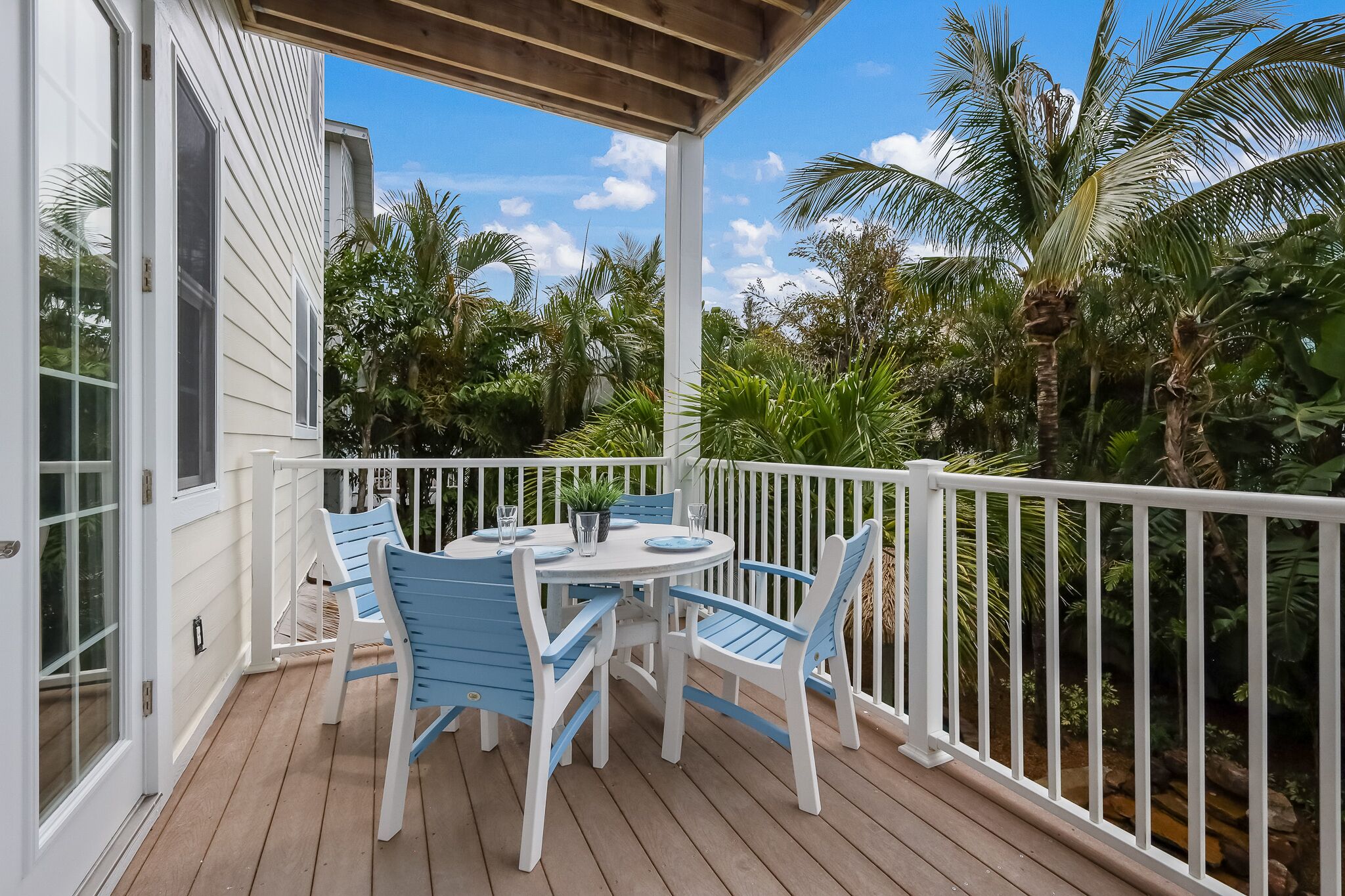 A covered outdoor area with a white railing, a round table with blue chairs, and a view of lush greenery.