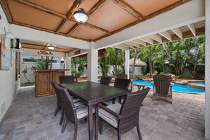 Patio esterno con tavolo e sedie, piscina e vegetazione lussureggiante.
