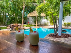 Deux verres contenant une boisson sont posés sur une table en bois, avec une piscine et des plantes tropicales en arrière-plan.