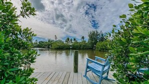 A wooden dock with a blue rocking chair, surrounded by lush greenery and a calm body of water. - Hammock Dreams - Canal Home with Pool and Dock Near Beach (Anna Maria)