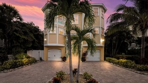 A multi-story building with a driveway leading to a garage, surrounded by palm trees and landscaped areas. - Poseidons Palace! Family Fun and Gulf Views! (Holmes Beach)