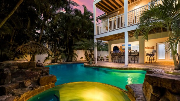 A pool area with two distinct sections, a covered seating area with tables and chairs, and a view of a building with a balcony. - Poseidons Palace! Family Fun and Gulf Views! (Holmes Beach)