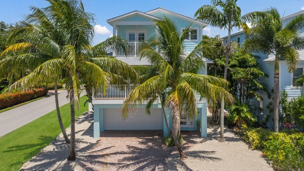 A two-story house with a light blue exterior, a balcony on the second floor, and surrounded by palm trees and tropical plants.