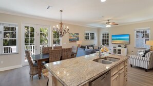 A spacious kitchen with a large island, dining area with wicker chairs, a living room with a sofa and TV, and large windows with a view.