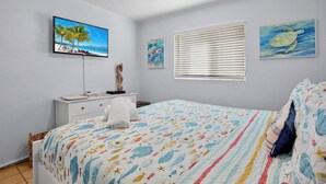 A bedroom with a bed, a flat-screen TV, a window with blinds, and artwork on the walls. - Beach Home with Pool Access and Private Patio with BBQ Grill! - Little Dream (Holmes Beach)