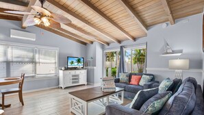 A spacious living room with a wooden ceiling, a large grey sofa, a coffee table, and a TV on a white cabinet.