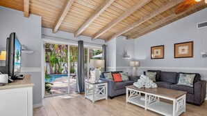 A living room with a grey sofa, white coffee table, and a TV on a stand. There is a sliding glass door leading to a pool area.