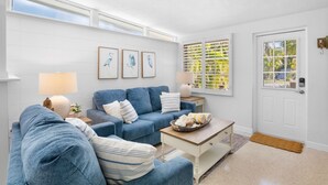 A living room with a blue sofa, a coffee table, and a lamp. There are framed pictures on the wall and a window with shutters.
