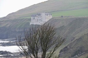 A large white building with multiple windows, situated on a hillside overlooking a body of water. - Bespoke 1 Bed Cottage in Dunbeath Village (Dunbeath)