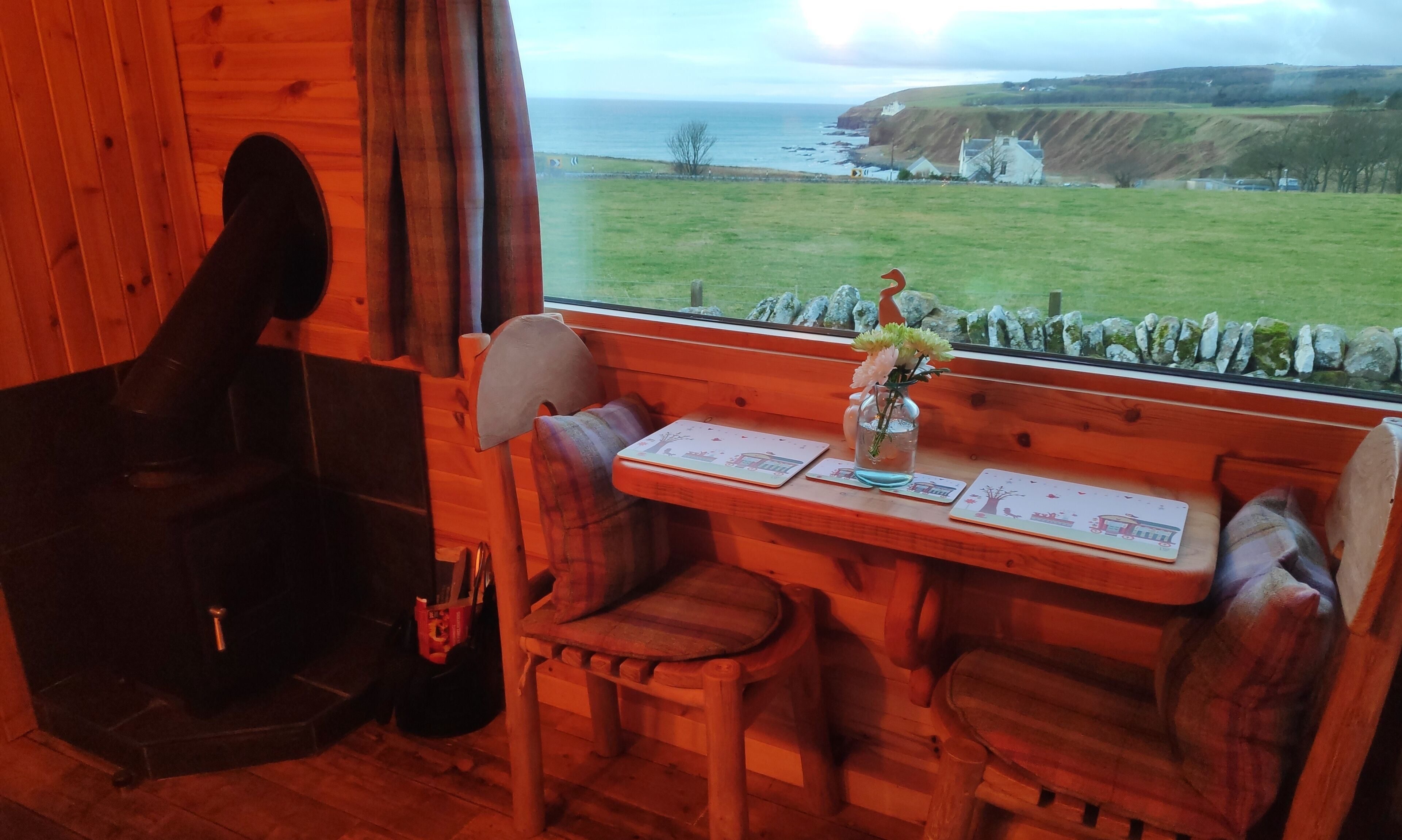 En mysig stuga med träinredning, ett bord med en vas med blommor och utsikt över havet och kullarna.